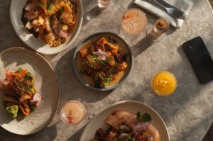 Overhead view of a restaurant table featuring various gourmet dishes and cocktails with dramatic sunlight and a smartphone placed on the side.