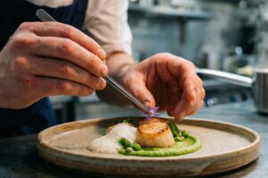 Close-up of a professional chef using tweezers to delicately garnish a gourmet scallop dish featuring green puree and foam.