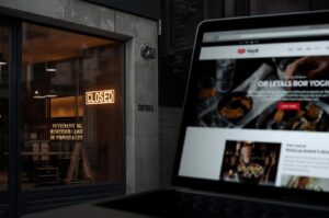 A view of a dark restaurant storefront with a glowing "Closed" neon sign, contrasted with a laptop in the foreground displaying an online food website or business listing.