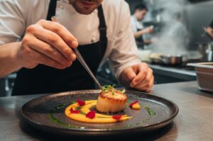 Professional chef in a commercial kitchen using tweezers to place a green garnish on a seared scallop dish featuring yellow purée and edible flower petals