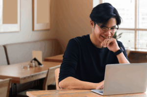 A young man with glasses sitting in a modern cafe, smiling while looking at his laptop screen. This illustrates the positive user experience of finding a great dining spot through online searches.