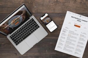 Top-down view of a wooden desk featuring a restaurant's branding across a laptop screen, a smartphone app, and a printed paper menu.