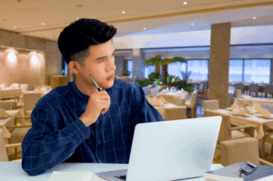 A pensive man sitting in a restaurant with a laptop, holding a pen to his chin and looking away thoughtfully. This depicts a restaurant owner concerned about why their business is not appearing in search results.