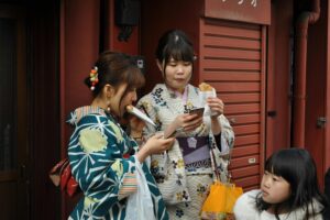 Two women wearing traditional Japanese kimonos stand together on a street, both eating snacks while focused on their smartphones.