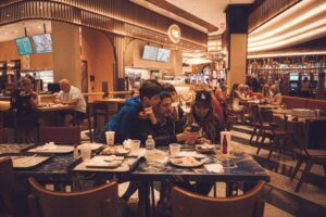 A family gathers closely around a smartphone at a blue marbled table in a busy, warm-lit food court. The surrounding area is filled with other diners and various food stalls.