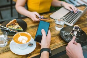 Two people sit at a wooden cafe table filled with coffee, water, and plates of food while using their laptops and smartphones.