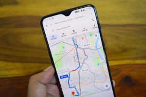 A hand holds a smartphone displaying Google Maps directions from NDLS railway station to Hauz Khas Lake.