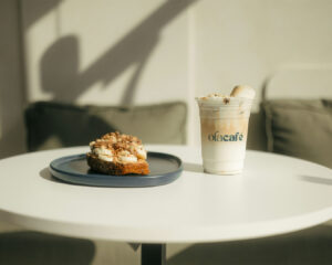 A slice of dessert served on a plate next to a cup of hot coffee on a café table, with soft natural light and blurred background creating a cozy, relaxed café atmosphere.