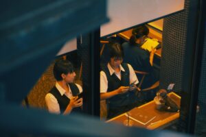 From an elevated perspective, two students in vests and white shirts are seen through a window using their smartphones at a cafe table.