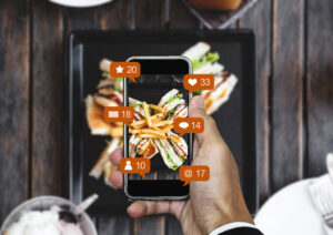 A hand holds a smartphone over a club sandwich and fries, capturing a photo for social media. Floating orange icons overlay the screen displaying interactions.