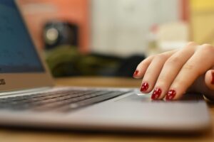 A hand with glossy red manicured nails rests on the trackpad of a silver laptop. The camera uses a shallow depth of field.