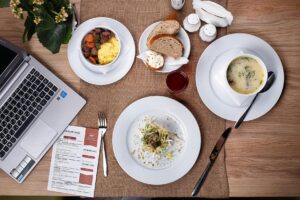 An overhead shot shows a multi-course meal arranged on a wooden table alongside a laptop and a printed menu.