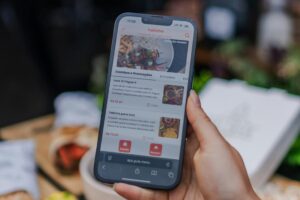 A person holds a smartphone displaying a digital food menu with various "Combos e Promocoes" listed in Portuguese. The background is softly blurred, showing hints of a dining area.