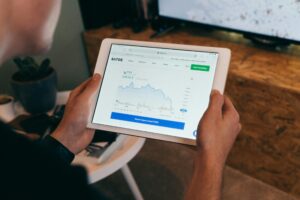 A person holds a white tablet displaying a financial website with a prominent line graph and the text "SATOS." They appear to be monitoring market trends or cryptocurrency prices while sitting in a dimly lit indoor setting.