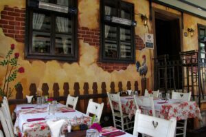 This charming outdoor restaurant patio features tables set with floral-patterned tablecloths and white wooden chairs with heart-shaped cutouts. The warm, rustic atmosphere is enhanced by a wall mural depicting roses, a rooster, and a picket fence.
