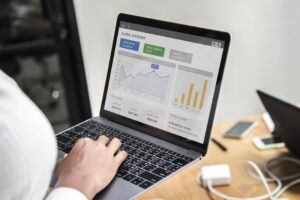 This image shows a person working on a laptop that displays a "Global Overview" dashboard featuring various financial charts and performance metrics. The scene is captured from an over-the-shoulder perspective on a wooden desk cluttered with tech accessories.