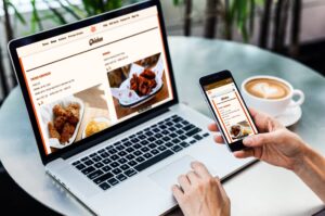 A person is using a laptop and a smartphone at a cafe table to view an online restaurant menu for "The Hot Chick." Beside the devices sits a cup of latte with heart-shaped latte art, emphasizing a modern, mobile dining experience.