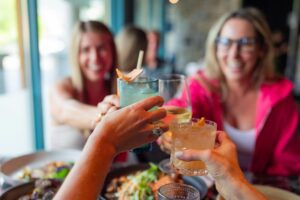 Three women are smiling and raising their glasses for a toast over a table of food. The foreground focuses on their hands holding colorful cocktails and wine, capturing a vibrant, social atmosphere.