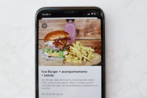 A close-up shot shows a smartphone displaying a food delivery app with a meal deal featuring a gourmet burger, french fries, and a pink bottled drink. The screen includes a Portuguese description of the "Sow Burger" along with its ingredients and a discounted price of R$ 35,00.