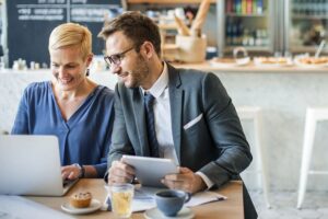 A man and a woman in professional attire are sitting at a cafe table, smiling while looking at a laptop and a tablet. The setting features a bright, modern interior with drinks and a small pastry on the table in front of them.