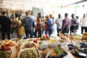 A large, diverse group of people gathers in a bright, modern space for a social event or party. In the foreground, a wooden table is spread with an abundant variety of catered dishes, including pasta, salads, and fresh seafood.