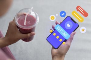 A person holds a purple smoothie in one hand while using their smartphone to leave a positive "AWESOME!" review with the other. Vibrant digital icons, including gold stars and speech bubbles, float around the screen to illustrate a successful customer experience.