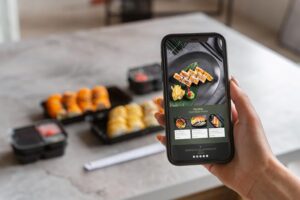 A person's hand holds a smartphone displaying a sushi restaurant's digital menu against a blurred background of a stone table. In the distance, several black takeout containers filled with various sushi rolls and condiments are ready to be enjoyed.