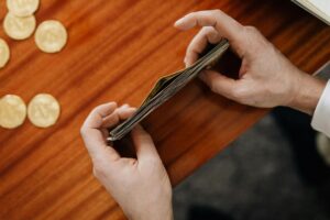 An overhead shot shows a person's hands holding and counting a thick stack of paper currency over a wooden surface. Several gold-colored coins are scattered to the left of the hands, suggesting a financial or budgeting context.