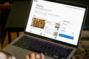 An open MacBook Pro displays the online ordering menu for a restaurant called Tony's Pizza, with a pizza named "Funghi Town" currently selected in the digital basket. A person's hand with light blue nail polish rests on the trackpad, seemingly ready to complete the checkout process.