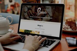 A person's hands are shown typing on a laptop that displays a restaurant website for "The Spoon," which features a "Serving a variety of fresh, homemade soup & pie daily" slogan. The laptop sits on a wooden table next to a white teapot, a smartphone, and a plate with a slice of cake.