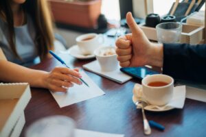 Two people sit at a wooden cafe table scattered with tea, snacks, and graph paper as one person gives a thumbs-up. The other person holds a blue pen over a notepad, appearing ready to jot down ideas during their brainstorming session.