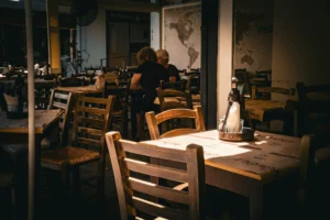 The image shows an empty restaurant patio filled with wooden tables and chairs, illuminated by a warm, dramatic shaft of sunlight hitting the foreground.