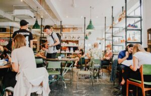 This image captures a bustling, industrial-style cafe filled with patrons sitting at green vintage chairs and wooden tables. Large open shelves displaying various goods and low-hanging green pendant lights create a bright, airy atmosphere throughout the space.