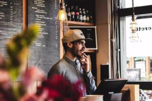 A man wearing a baseball cap and a denim shirt stands behind a cafe counter, looking thoughtfully toward a window. He is surrounded by industrial-style lighting and a large menu board listing various food and drink options.