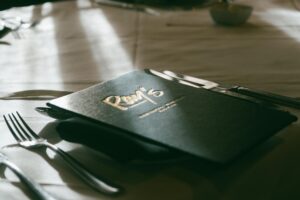 A black menu featuring the name "Ray's" in gold script rests on a table set for a meal. The scene is illuminated by soft, natural light, highlighting the textures of the tablecloth and silver cutlery.