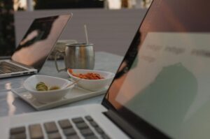 Two open laptops sit on an outdoor table alongside small bowls of olives and nuts. A metal mug with a straw is positioned between the computers.