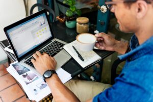 A man in a blue shirt works on a laptop at a cafe table while holding a cup of latte. His workspace is filled with professional tools, including a notebook, printed documents, and a small potted plant.