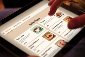 A person uses their fingers to scroll through a digital restaurant menu displayed on a tablet. The interface features a section titled "Italian Dolce" with photos and descriptions of desserts like Panna cotta and Tiramisu.