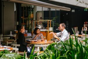 A small group of people are engaged in an animated conversation while dining at an upscale outdoor patio. Lush green plants frame the foreground, while the restaurant’s modern architecture and warm sunlight create a sophisticated atmosphere.