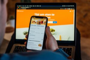 A person holds a smartphone displaying a McDonald's menu on a food delivery app, positioned in front of a laptop with a matching orange website. The scene captures a first-person perspective.
