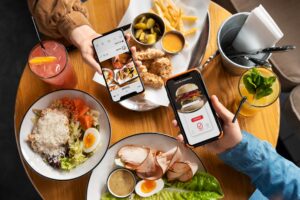 Two people are holding up their smartphones to take photos of a variety of dishes on a wooden table, including salads, fried chicken, and colorful drinks. The overhead shot captures the modern social dining experience, highlighting the interaction between technology and food.