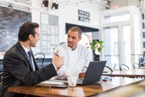 A businessman in a suit and a chef in a white coat are sitting together at a restaurant table, engaged in a focused discussion. They are using a laptop and documents to go over details in a bright, modern dining area.