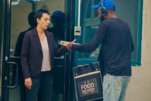 A delivery courier wearing a blue hat and headphones hands a pizza box to a woman at the entrance of a building. The courier carries a black insulated "FOOD DELIVERY" bag as the customer steps out to receive her meal.