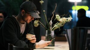 A young man wearing a baseball cap and glasses sits at a wooden table, holding a ceramic cup while looking at his smartphone. Beside him, a glass vase filled with flowers adds a touch of nature to the dimly lit, urban cafe setting.
