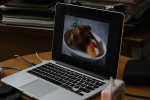 A silver laptop sits open on a wooden desk, displaying a photo of a chocolate-drizzled croissant and a cream-filled pastry on its screen. The surrounding workspace is cluttered with charging cables, electronic accessories, and a stack of notebooks in the background.