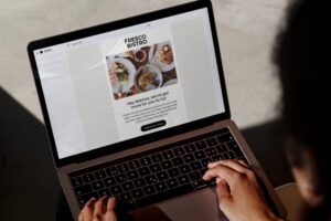A person is using a MacBook Pro to view a promotional email from Fresco Bistro featuring a photo of plated gourmet dishes. Their hands are positioned on the keyboard and trackpad while they browse the "Explore our menu" call to action.