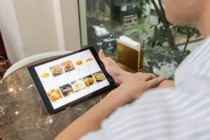 A person in a striped shirt sits at a marble table while holding a tablet displaying a digital food menu. The screen shows various appetizers like french fries and onion rings, with a street view visible through the window in the background.