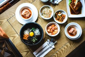 This image showcases a vibrant spread of Korean cuisine, featuring a central bowl of bibimbap topped with a bright egg yolk and various colorful vegetables. Surrounding the main dish are several small bowls of banchan, including kimchi, sprouts, and cucumbers, all neatly arranged on a light wooden table.