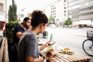 A woman with her hair in a bun sits at an outdoor cafe table, looking at her smartphone while preparing to eat a meal. In the background, a man sits at the same wooden table against a blurred backdrop of a city street with buildings and a passing bicycle.