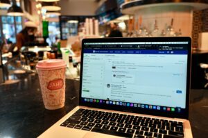 A MacBook Air sits on a dark cafe counter next to a pink "Jaho" coffee cup, displaying a LinkedIn Sales Navigator search and a low battery notification. In the background, the warm, blurred interior of a coffee shop shows staff and patrons in a cozy urban atmosphere.